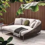 “Patio rope daybed set in dark brown frame with cream cushions, placed outdoors on a wooden deck surrounded by greenery.”