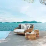 Beige and off-white outdoor rope daybed with plush cushions on a wooden deck surrounded by greenery.