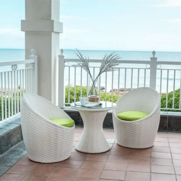 White 3-piece rattan wicker patio set with two chairs and a glass-top table on a balcony overlooking the sea.