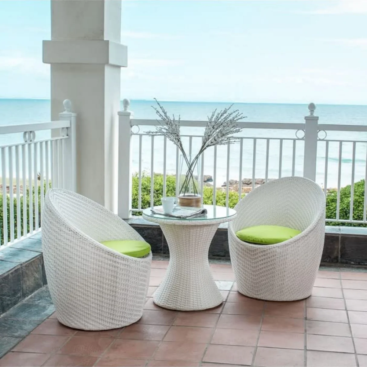 White 3-piece rattan wicker patio set with two chairs and a glass-top table on a balcony overlooking the sea.
