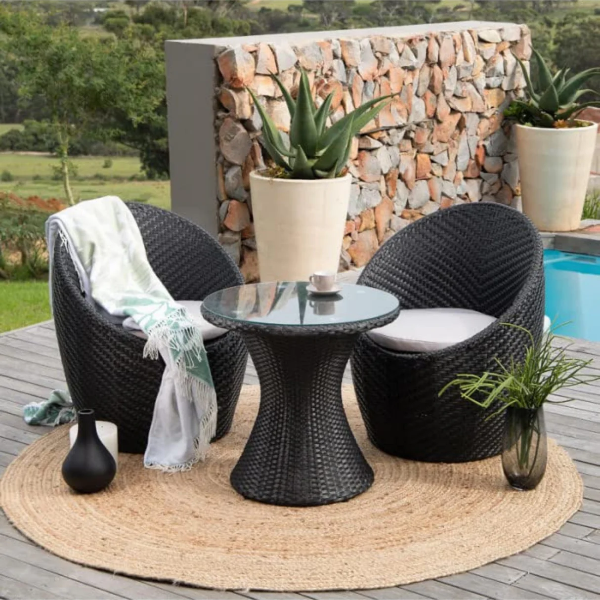 3-piece black rattan wicker patio set with glass-top table and cushioned chairs placed on a round jute rug beside a pool.