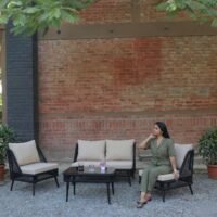 Outdoor patio furniture setup with a black woven sofa set, including a two-seater, two single chairs, and a coffee table with drinks and a small flower vase, placed on a gravel surface against a red brick wall. A woman in a green outfit is seated on one chair, relaxing under leafy trees in a garden setting.