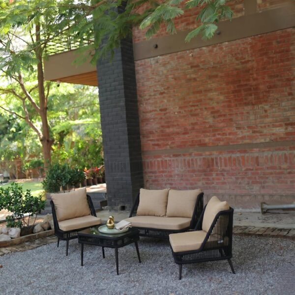 Outdoor patio furniture setup with a black woven sofa set, including a two-seater, two single chairs, and a coffee table with drinks and a small flower vase, placed on a gravel surface against a red brick wall. A woman in a green outfit is seated on one chair, relaxing under leafy trees in a garden setting.