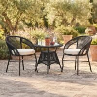 3-piece outdoor rope patio furniture set with two black handwoven rope chairs, cream cushions, and a round matching table in a garden patio setting.
